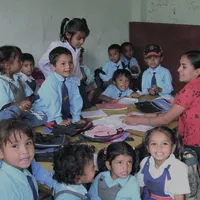 A group of young children in school uniforms gather around a table with their teacher in a classroom setting, smiling and engaged.