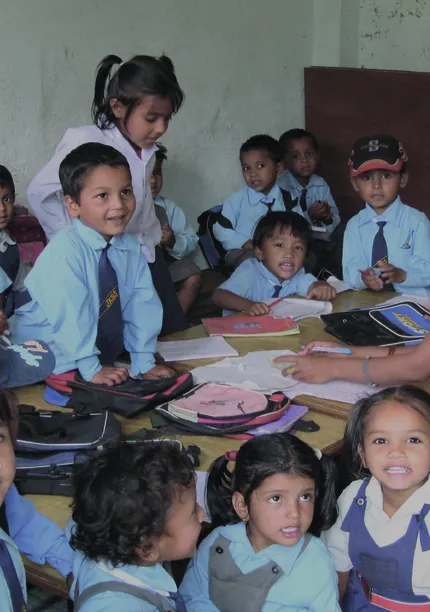 A group of young children in school uniforms gather around a table with their teacher in a classroom setting, smiling and engaged.