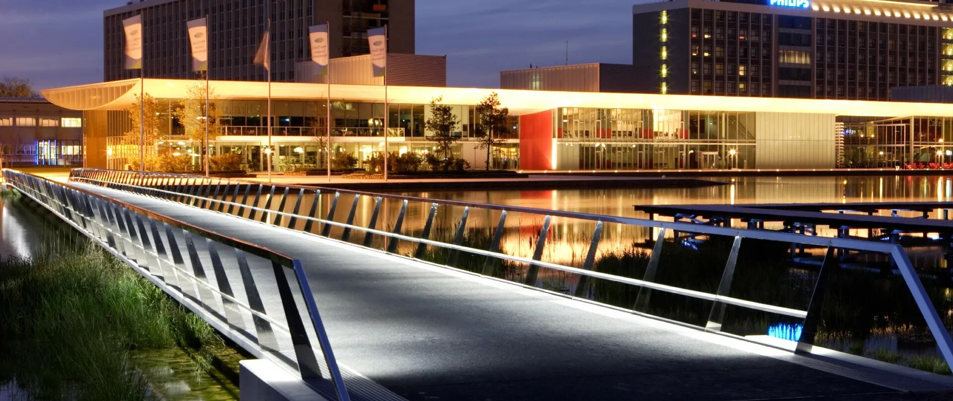 Illuminated modern bridge leading to buildings at dusk, with reflections on the water. Buildings have visible signage and warm lighting.