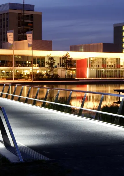Illuminated modern bridge leading to buildings at dusk, with reflections on the water. Buildings have visible signage and warm lighting.