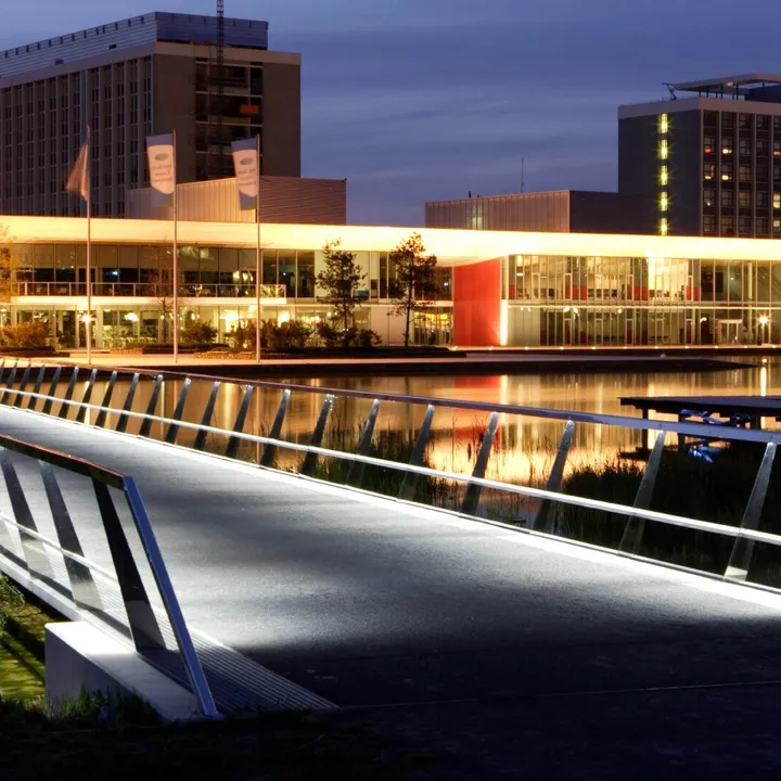 Illuminated modern bridge leading to buildings at dusk, with reflections on the water. Buildings have visible signage and warm lighting.