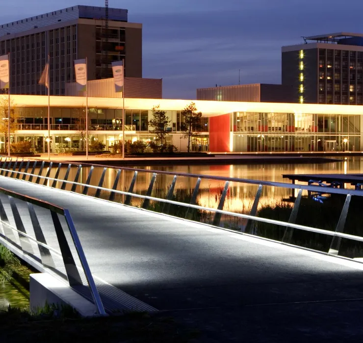 Illuminated modern bridge leading to buildings at dusk, with reflections on the water. Buildings have visible signage and warm lighting.