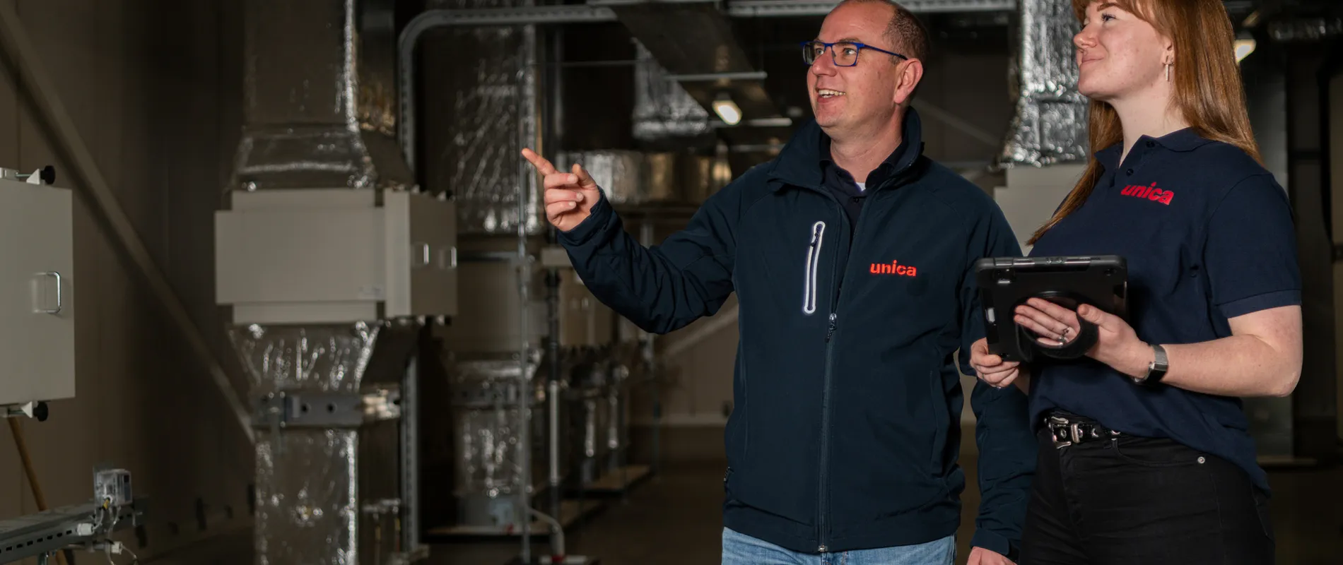 "Twee medewerkers van Unica Watertechniek inspecteren een technische ruimte met HVAC-installaties, gericht op efficiënte en veilige wateroplossingen."
