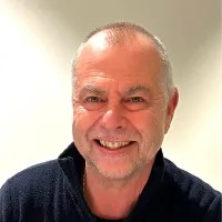 Smiling older man with short hair and a grey beard, wearing a dark zip-up sweater, against a neutral background.