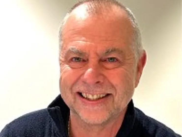 Smiling older man with short hair and a grey beard, wearing a dark zip-up sweater, against a neutral background.