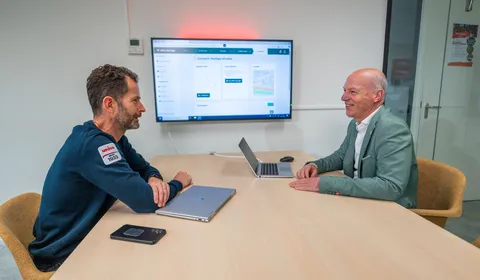 Twee collega’s in gesprek met op de achtergrond het EMS-platform geprojecteerd op een scherm.