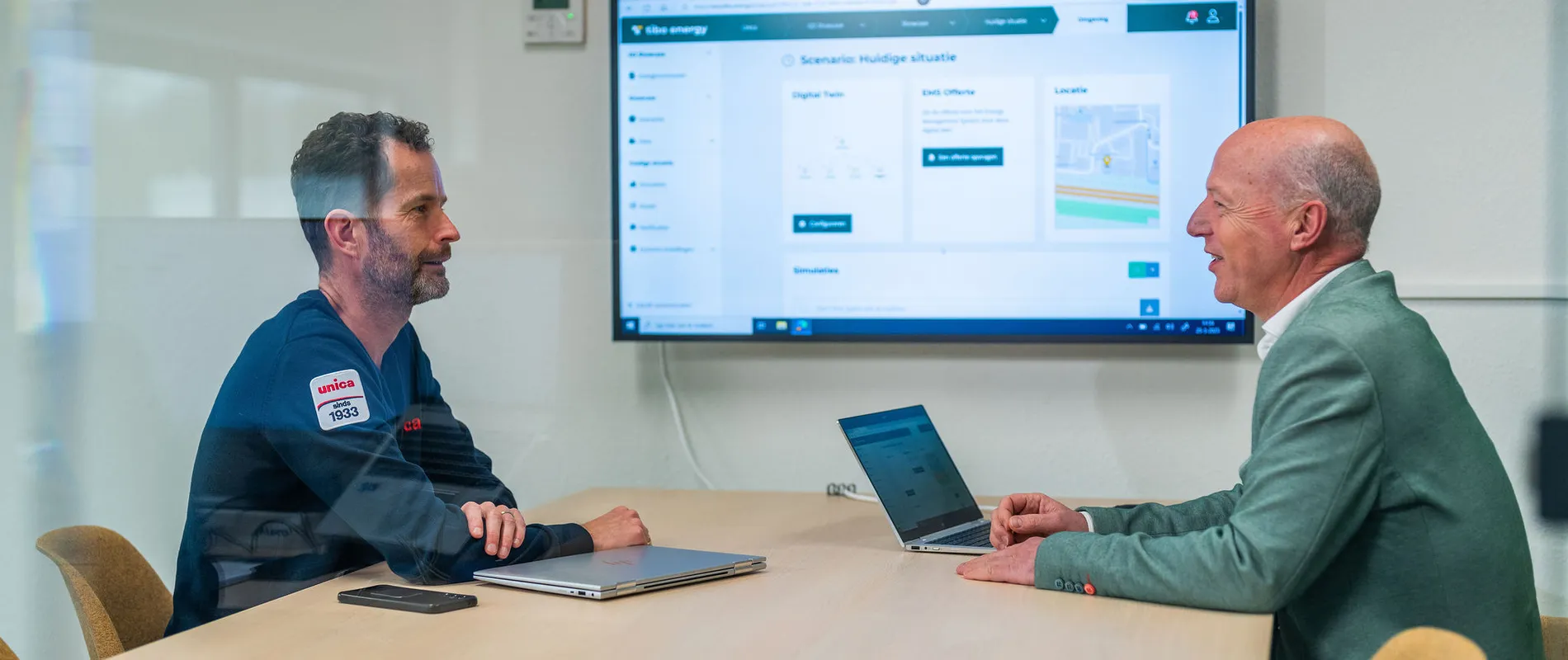 Twee mannen in overleg bij Unica over energiemanagementsystemen met een dashboard op het scherm.