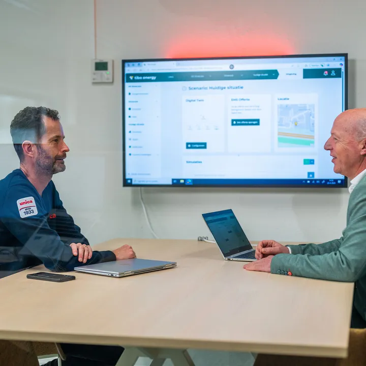 Twee mannen in overleg bij Unica over energiemanagementsystemen met een dashboard op het scherm.