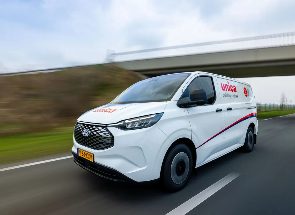 White Unica van with logo driving on a highway, under an overpass, on a clear day. License plate visible, road and sky in the background.