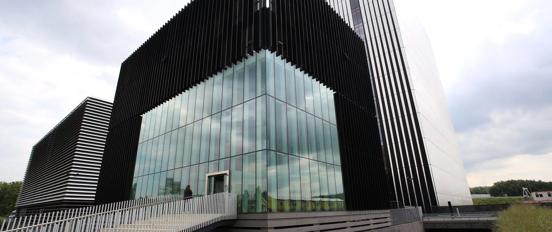 Modern building with black vertical slats and large glass windows, featuring a sloped white wall and a bridge over a reflective pond.
