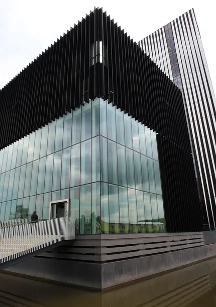 Modern building with black vertical slats and large glass windows, featuring a sloped white wall and a bridge over a reflective pond.