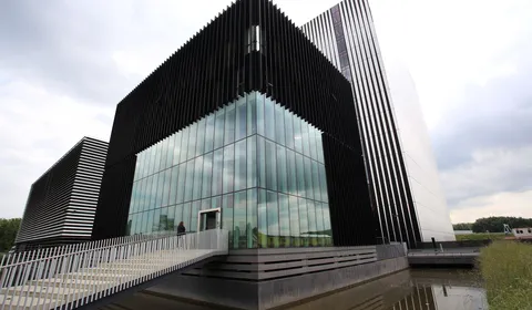 Modern building with black vertical slats and large glass windows, featuring a sloped white wall and a bridge over a reflective pond.