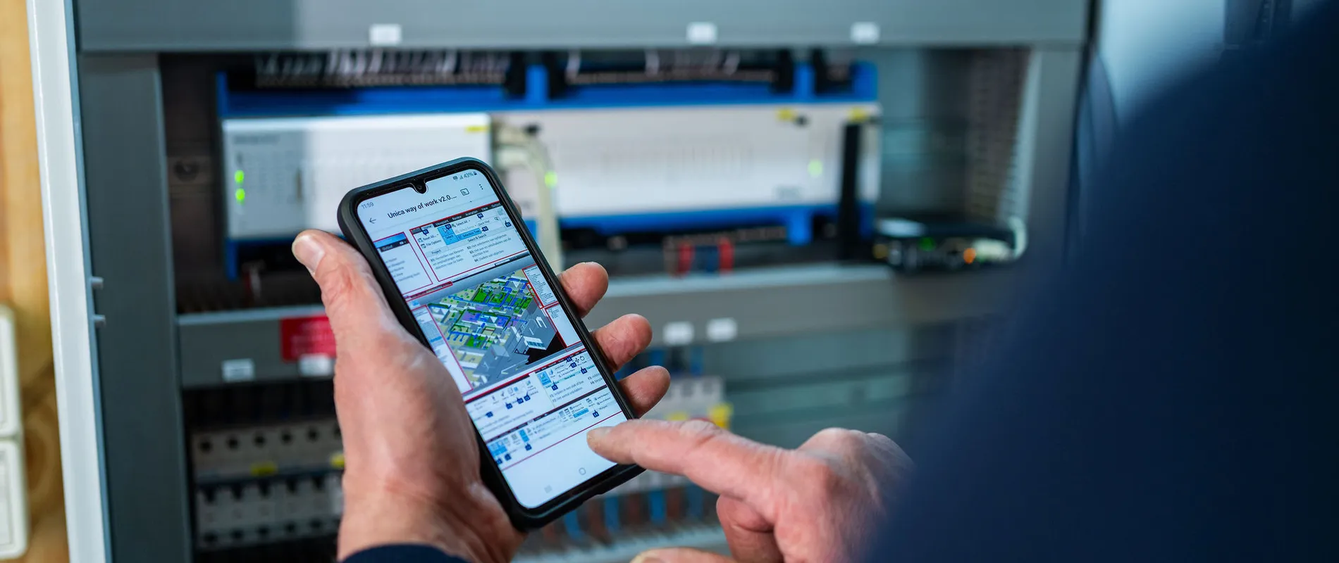 Unica technicus gebruikt smartphone om elektrotechnische installatie te monitoren in schakelkast bij gebouwbeheer.