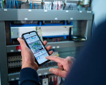 Unica technicus gebruikt smartphone om elektrotechnische installatie te monitoren in schakelkast bij gebouwbeheer.