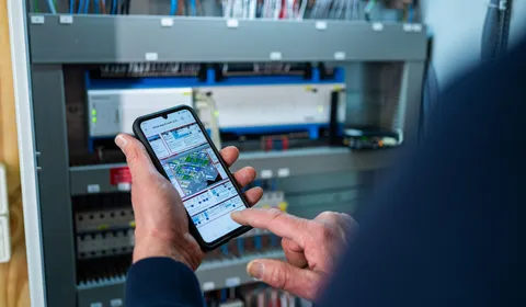 Unica technicus gebruikt smartphone om elektrotechnische installatie te monitoren in schakelkast bij gebouwbeheer.