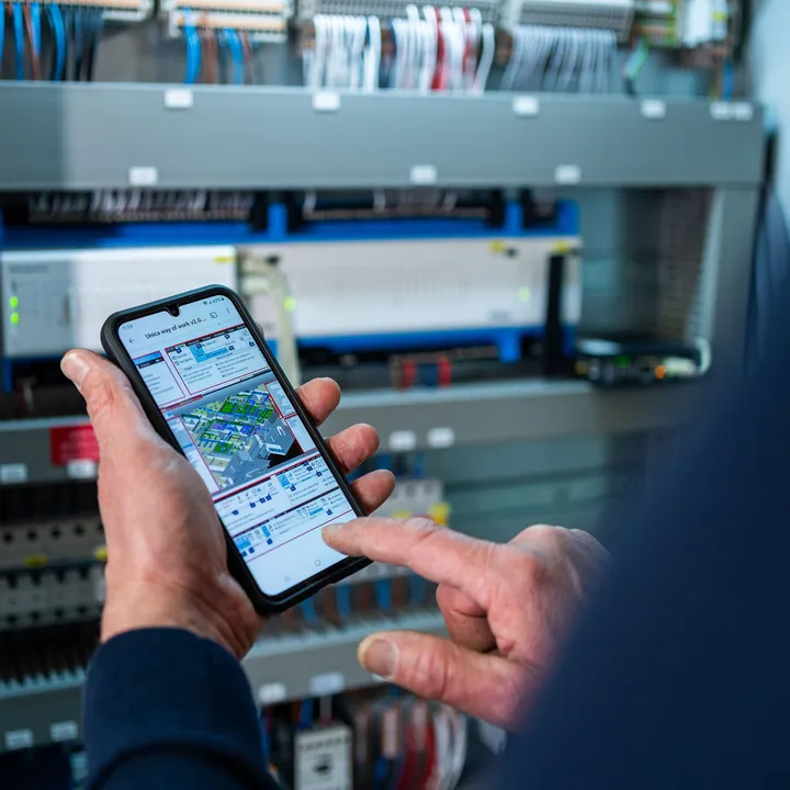 Unica technicus gebruikt smartphone om elektrotechnische installatie te monitoren in schakelkast bij gebouwbeheer.
