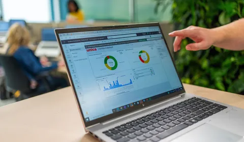 Een medewerker wijst naar een digitaal dashboard met klanttevredenheidsscores op een laptop, met op de achtergrond collega’s in een moderne kantooromgeving.