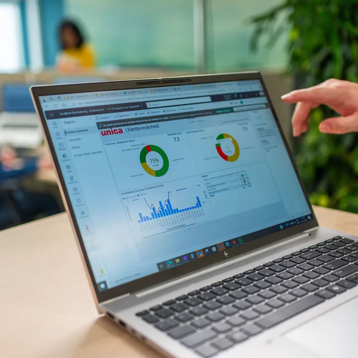 Een medewerker wijst naar een digitaal dashboard met klanttevredenheidsscores op een laptop, met op de achtergrond collega’s in een moderne kantooromgeving.