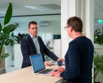 Twee collega’s bespreken verduurzamingsopties met laptops op een sta-bureau in een licht kantoor met planten.