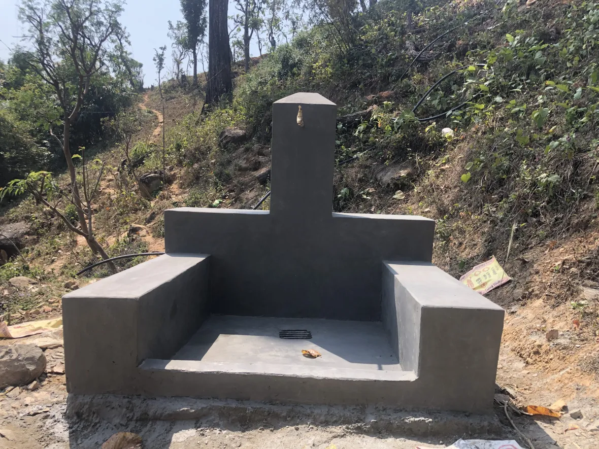 Concrete water tap structure in a rural, hilly area, with pipes extending from the back and surrounded by trees and vegetation.