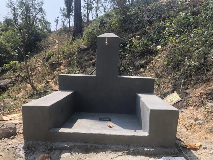 Concrete water tap structure in a rural, hilly area, with pipes extending from the back and surrounded by trees and vegetation.