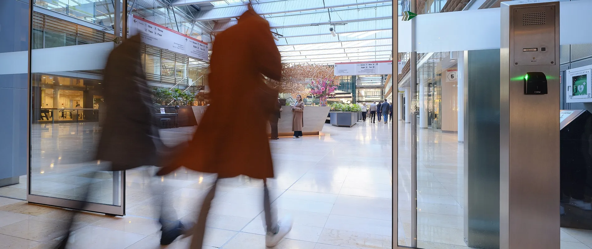 Two people walking briskly through a modern, glass-roofed atrium with shops and greenery, creating a motion blur effect.