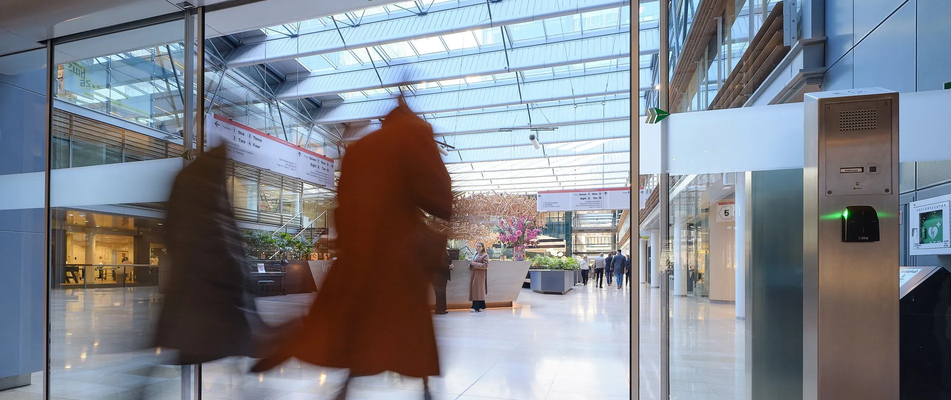 Two people walking briskly through a modern, glass-roofed atrium with shops and greenery, creating a motion blur effect.