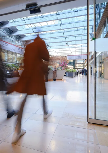 Two people walking briskly through a modern, glass-roofed atrium with shops and greenery, creating a motion blur effect.
