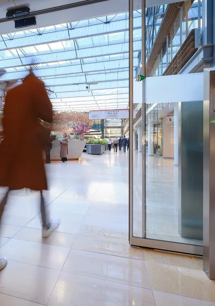 Two people walking briskly through a modern, glass-roofed atrium with shops and greenery, creating a motion blur effect.