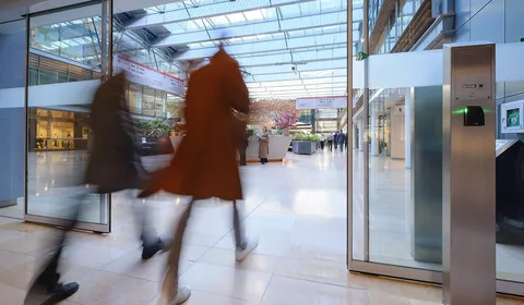 Two people walking briskly through a modern, glass-roofed atrium with shops and greenery, creating a motion blur effect.