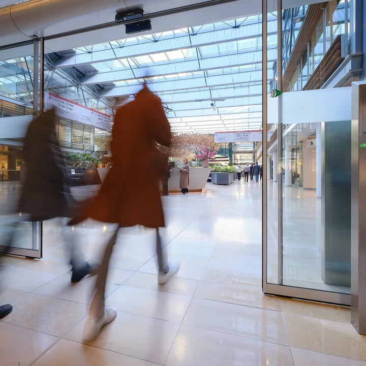 Two people walking briskly through a modern, glass-roofed atrium with shops and greenery, creating a motion blur effect.