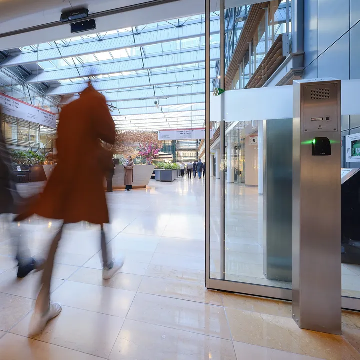 Two people walking briskly through a modern, glass-roofed atrium with shops and greenery, creating a motion blur effect.