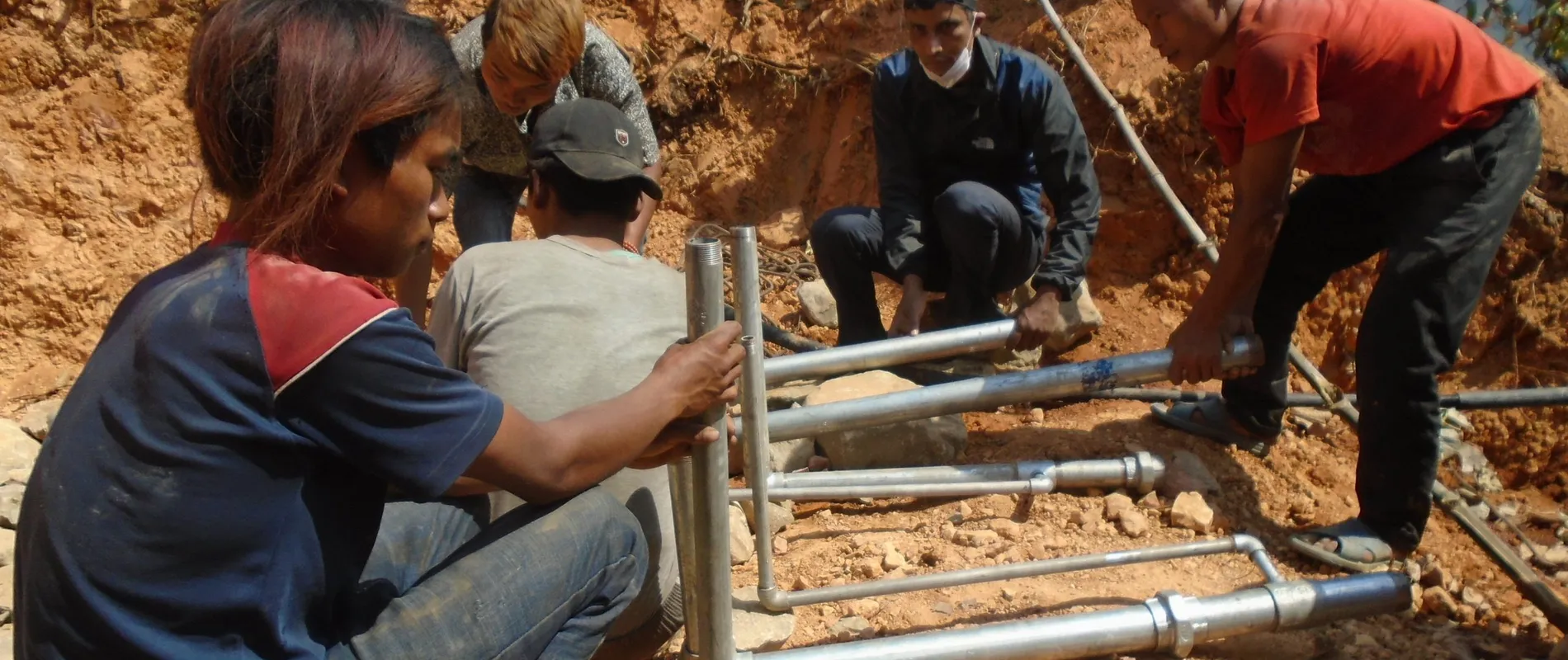 People in a rural setting are assembling a metal structure on a dirt ground, surrounded by rocks and vegetation.
