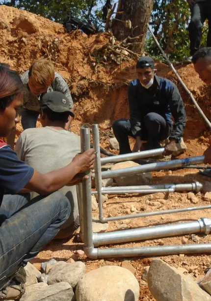 People in a rural setting are assembling a metal structure on a dirt ground, surrounded by rocks and vegetation.