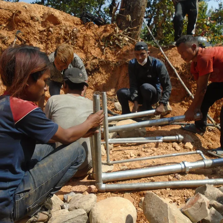 People in a rural setting are assembling a metal structure on a dirt ground, surrounded by rocks and vegetation.