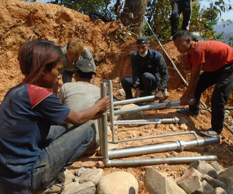 People assembling metal pipes at a construction site with dirt and rocks, under sunny conditions.