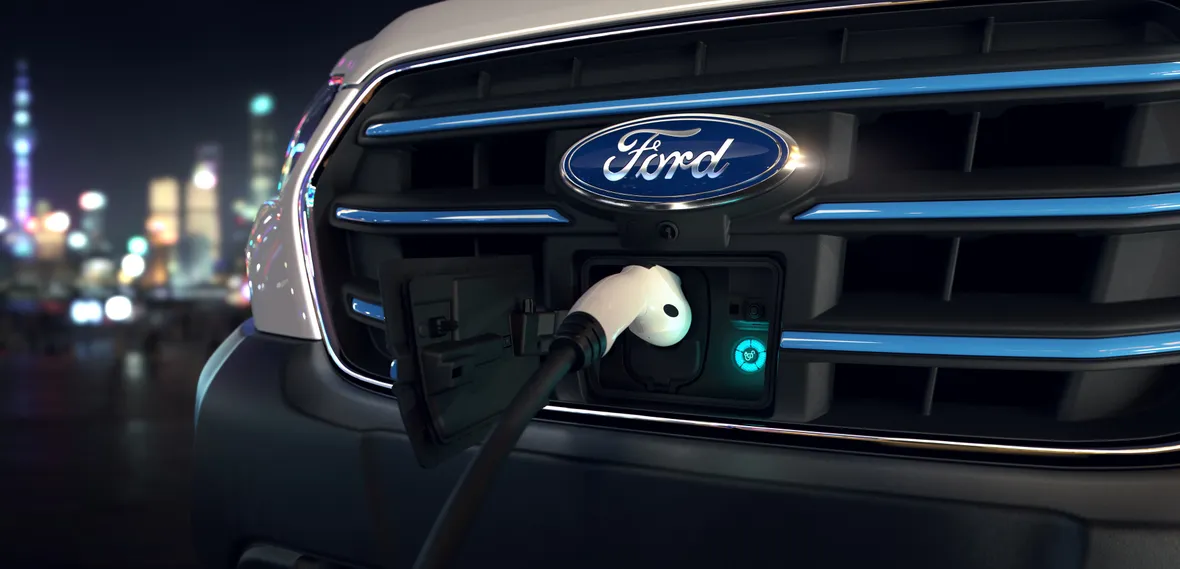 Close-up of a Ford vehicle's front grille with an electric charging cable connected, city lights blurred in the background.