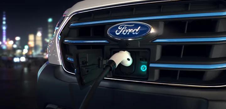 Close-up of a Ford vehicle's front grille with an electric charging cable connected, city lights blurred in the background.