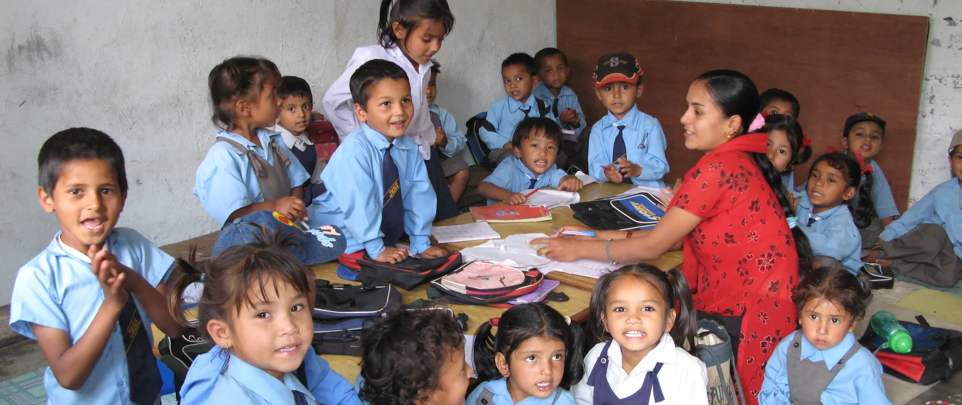 A teacher sits with a group of young children in blue uniforms, smiling in a classroom with educational materials on the table.