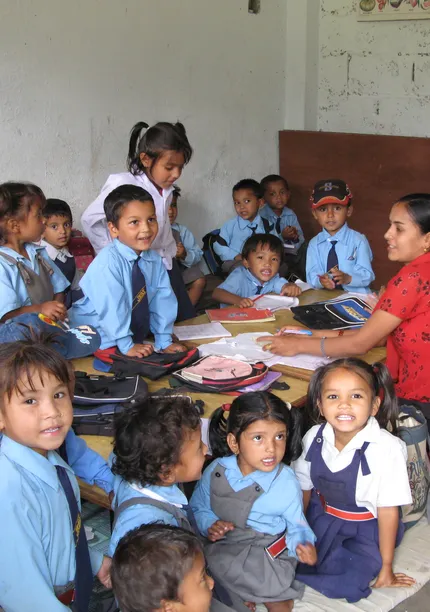 A teacher sits with a group of young children in blue uniforms, smiling in a classroom with educational materials on the table.