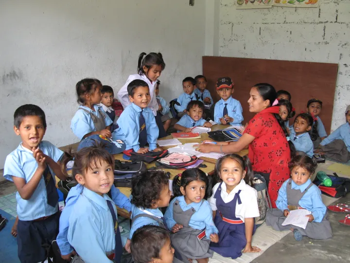 A teacher sits with a group of young children in blue uniforms, smiling in a classroom with educational materials on the table.