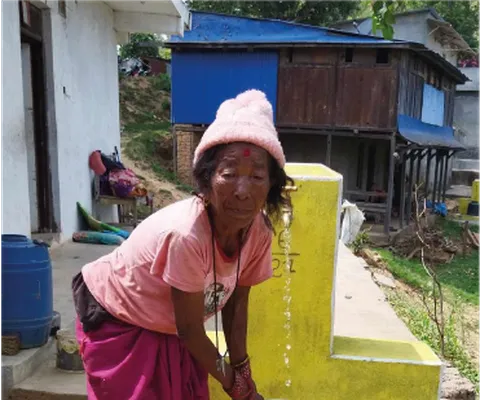 An older woman fills a metal water jug from a public tap in a rural setting, wearing a pink hat, shirt, and red skirt.