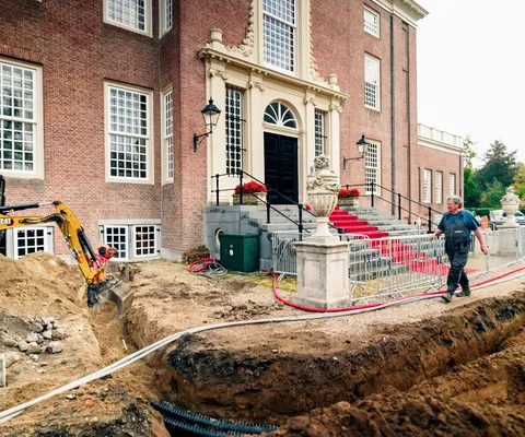 Construction workers dig trenches near a historic brick building with a red carpet entrance, using a mini excavator and laying pipes.
