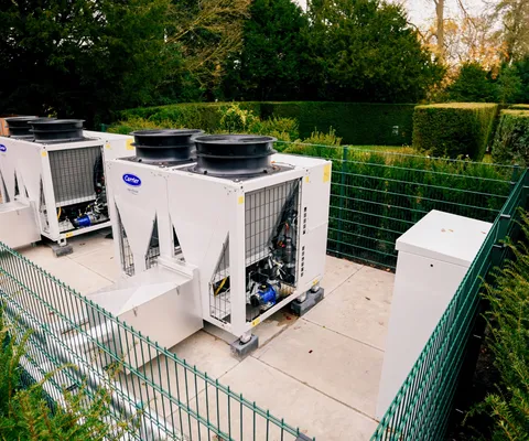 Three industrial HVAC units are enclosed by a green metal fence, surrounded by greenery on a concrete platform outdoors.