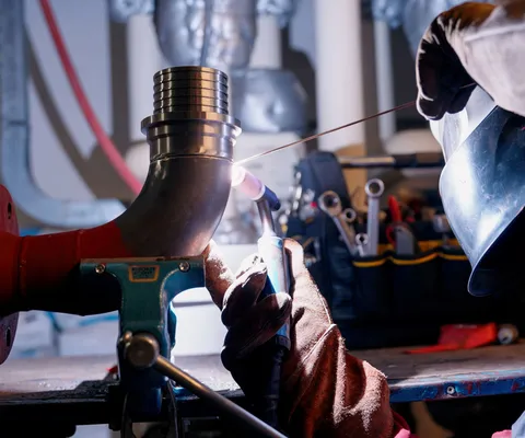 Person welding a metal pipe on a workbench, wearing protective gloves and helmet, with tools visible in the background.