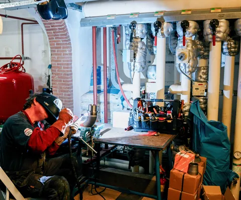A person wearing protective gear welding in a mechanical room, surrounded by insulated pipes, tools, a red tank, and a brick archway.