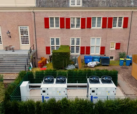 Aerial view of a brick building with red shutters, steps, outdoor seating, and adjacent fenced-in HVAC units surrounded by greenery.