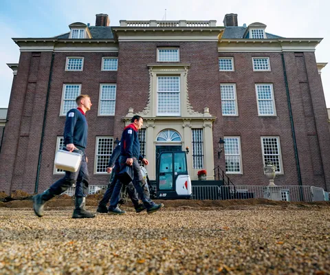 Three workers in uniforms walk in front of a large, historic brick building under a clear sky.