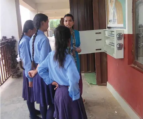 Four students in blue uniforms listen to a teacher outside a school building, near a wall painting of Mother Teresa.