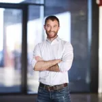 Smiling man with a beard, wearing a white shirt and jeans, stands with arms crossed in a modern, well-lit interior with large windows.